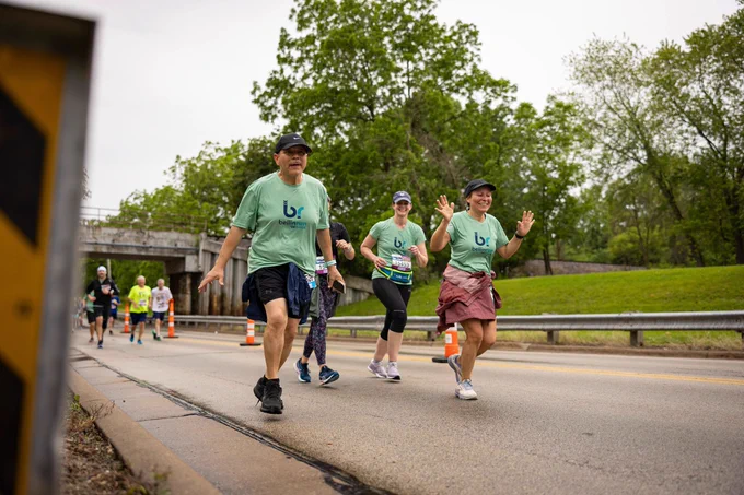 Group of runners wearing matching green shirts participating in a road race event, with a bridge and trees in the background.