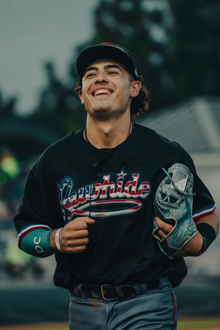 Demetrio Crisantes smiles while wearing a black Visalia Rawhide uniform top with the team's script in U.S. flag colors.