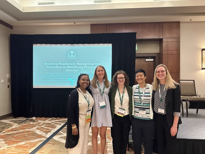 Five people stand together in front of a projector screen and short stage, all smiling at the camera. The projector screen behind them reads "Building Resilience: Navigating the Transition to Residency Program Director".