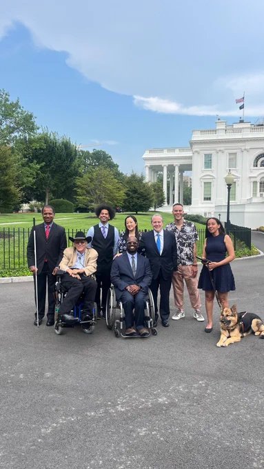 Disability advocates, including many people of color, smile in front of the White House. From left to right: Dr. Sachin Pavithran, Kelly Buckland, Mychal Threets, Eric Harris, Anna Perng, Gene Sperling, James Ian, Haben Girma, and her guide dog Mylo. Behind them is a long paved road that stretches in front of the West Wing. Photo by @WhiteHouseOPE46