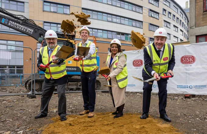 A picture of Ben Travis (LGT CEO), Mayor of Lewisham Brenda Dacres and representatives from Sisk (construction partners) tossing soil into the air with shovels, in the construction site of Lewisham Surgical Centre. All are wearing hard hats and hi-vis jackets.