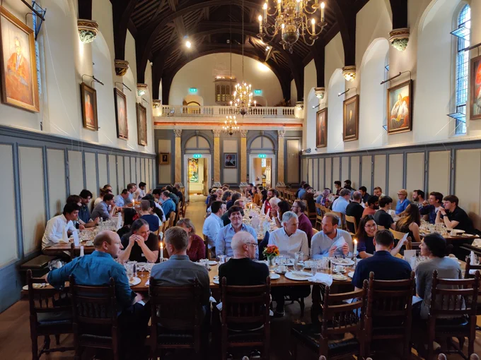 Photograph of the inside of a large dining hall with blue and cream panelled walls and a dark wood vaulted ceiling. Four tables are full of people enjoying food and conversation!