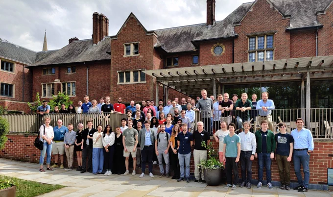 A photograph of a large group of people standing on and around some steps leading up to a terrace in front of a red brick building. 