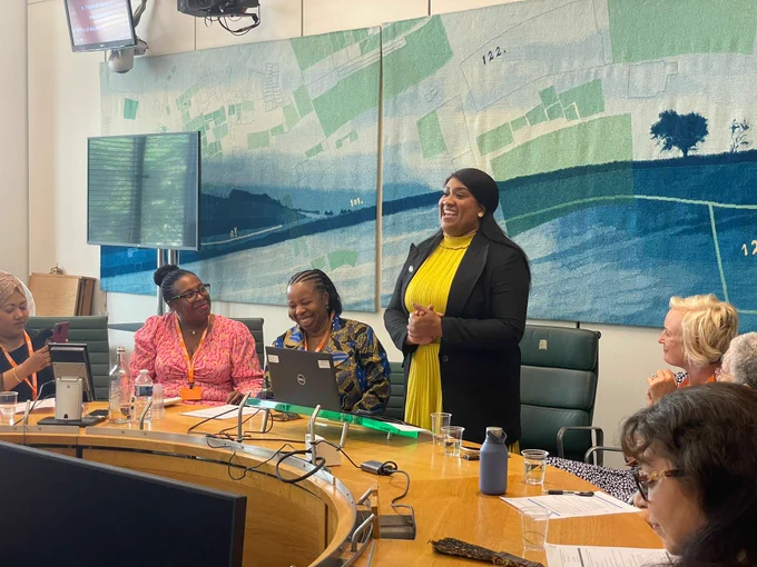 Bell speaks from a parliamentary roundtable with several women sitting around her. There are microphones, water glasses and notes on the table in front of them.