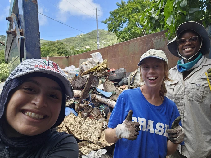 A selfie of three people smiling standing in front of a dumpster with collected garbage.