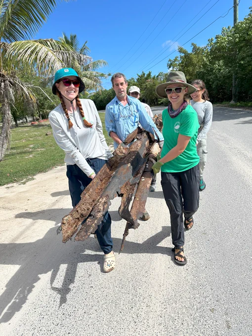 Six people carry a large, rusted metal beam down the road.
