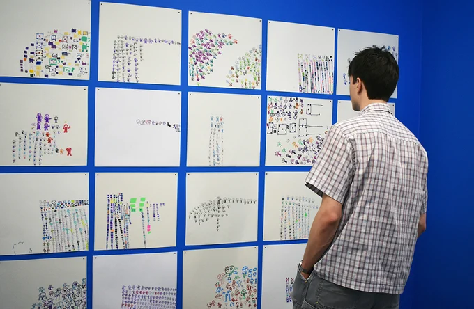 Photo of a person looking at a series of drawings arranged in a grid form on a blue wall. 