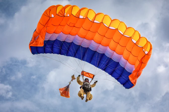 A smokejumper is seen from below as he dangles beneath a brightly colored parachute. He is looking down and pulling with both hands on the steering lines. The blue, lavender, orange, and yellow of the parachute are vivid against a blue sky with thin white clouds. Photo by Heal Herbert, DOI.