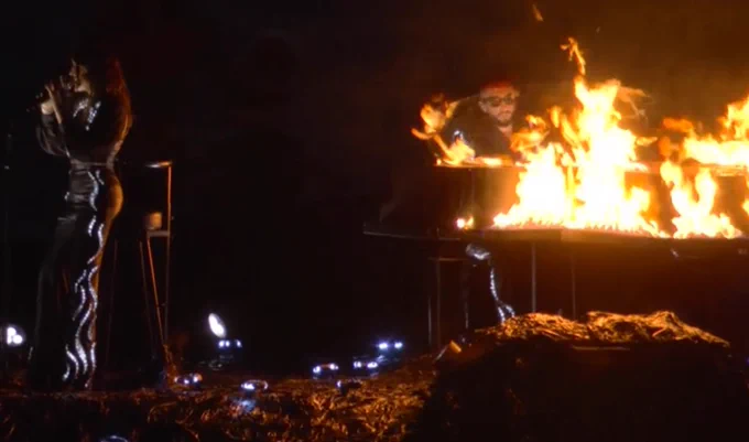 A woman performing next to a man next a piano on fire