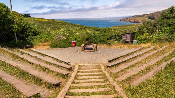 An outdoor ampitheatre with wooden benches and a firepit. Green hills and blue ocean are in the background.