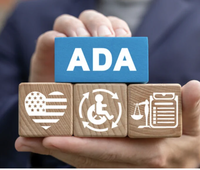 Three wooden blocks with the word ADA on the top block, an American flag heart on the left block, a symbol of a person in a wheelchair on the middle block, and a scale and document on the right block.