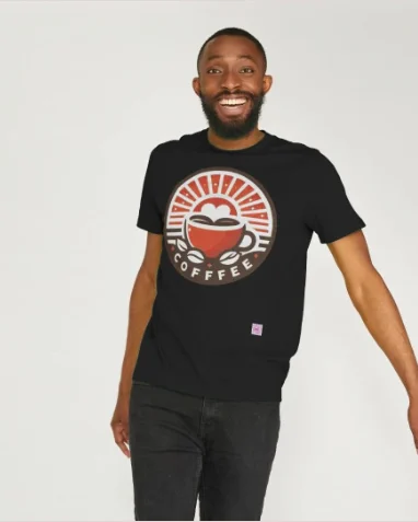 A unisex T-shirt black material with large chest circular logo of a sunrise in orange beams on a white background over a white heart sitting in a coffee cup in orange with half cut coffee beans left & right of the base of the cup, below this is the word coffee in white capital letters the image has a brown border, In the bottom corner is the QR code of Klazva.com in 3 cm square.This has a man wearing it. Available at Klazva.teemill.com main site Klazva.com.