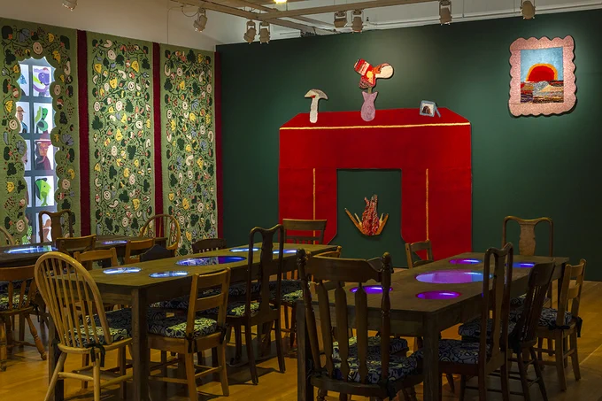 A photograph of a dimly lit room with three wooden tables surrounded by chairs. The far wall is dark green with a large artwork resembling a red fireplace and framed picture. On the left wall is a lighter green tapestry with a busy floral pattern.