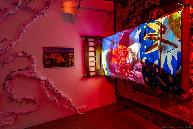 An installation view of a corner of a red-lit room with a bed, rug, and upright lamp installed on the wall to the right. On the wall to the left is a poster of the film, Psycho, with long paper coils that extend from the floor to the ceiling.