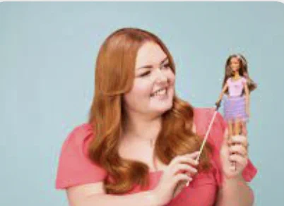 A woman with long red hair is smiling and holding a Barbie doll with a white stick.
