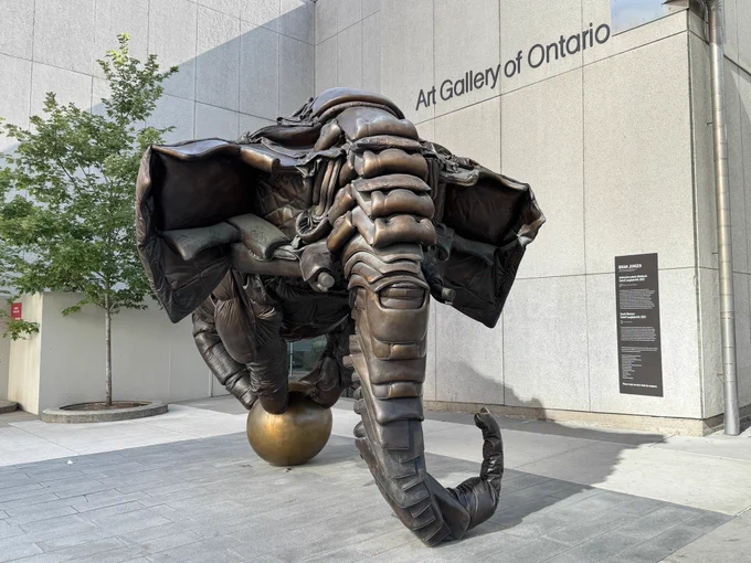 A metal sculptor of a stylized elephant balancing on a ball, with the exterior of the Art Gallery of Ontario and a small tree in the background. The artist’s name is Sadzěʔ yaaghęhch’ill.