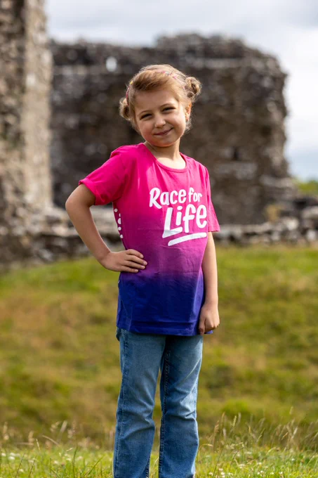 Betsy standing in a park, smiling with her hand on her hip. She is wearing a Race for Life t-shirt and jeans.