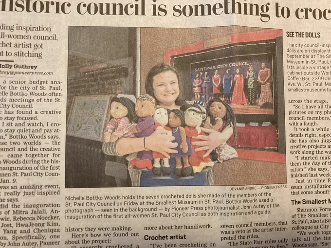 Newspaper photo of a woman holding 7 crocheted woman dolls in front of a photo of the same women as people. Colorful clothes.