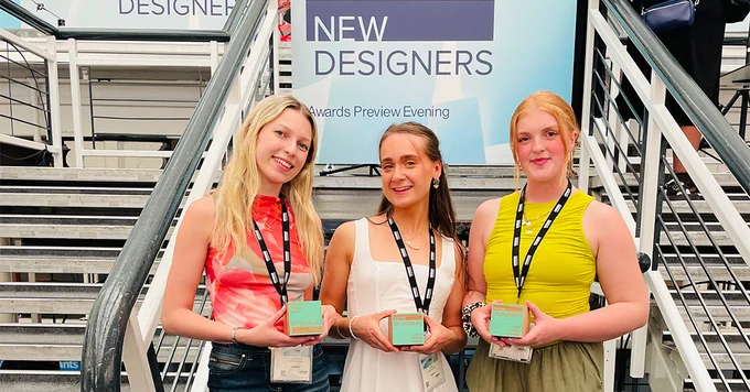 Charlotte Devereux, Ellie Munro and Amy Gardner with their New Designers Awards, smiling in front of a New Designer poster and a stair case