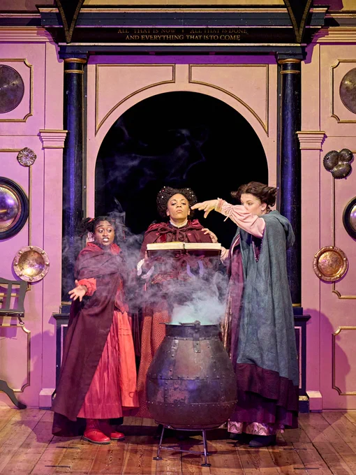 The company of Rough Magic (2024) onstage in the Sam Wanamaker Playhouse - stood in cloaks around a cauldron which has smoke coming out of it.