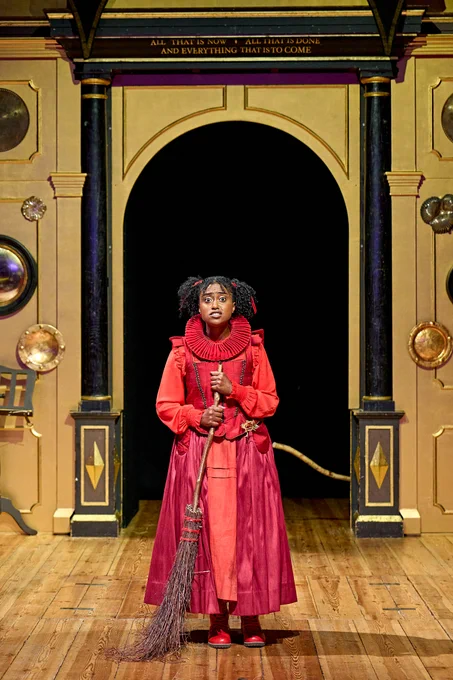The company of Rough Magic (2024) onstage in the Sam Wanamaker Playhouse - Rosemarie Akwafo stood in the centre in an ornate red costume, holding a broomstick and looking into the audience.