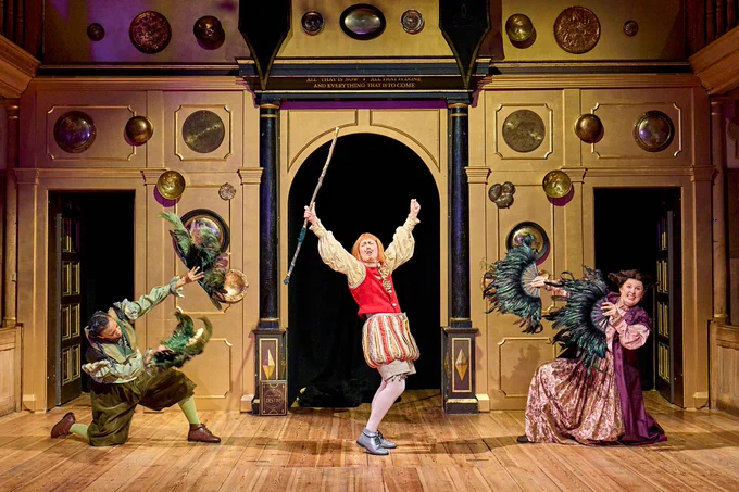 The company of Rough Magic (2024) onstage in the Sam Wanamaker Playhouse - in ornate Elizabethan style clothes. Kerry Frampton in the centre, arms up in the air triumphantly holding a staff, as other company members hold and wave fans.