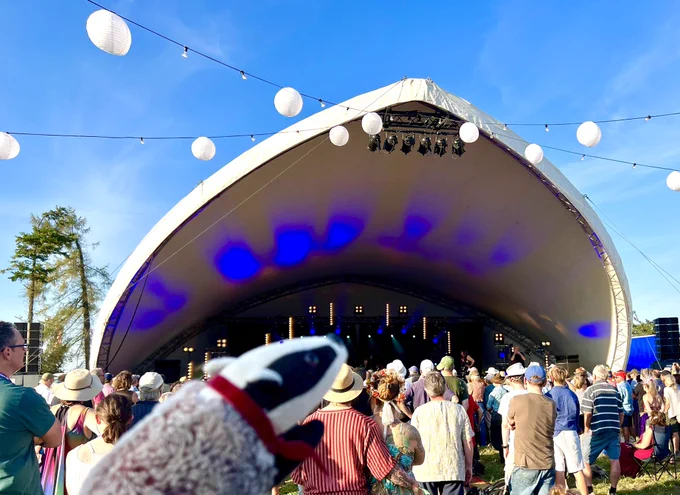 The parabolic roof of the Charlie Gillett stage against a bright blue sky, with back view of enthusiastic crowd. Small toy badger in left foreground.