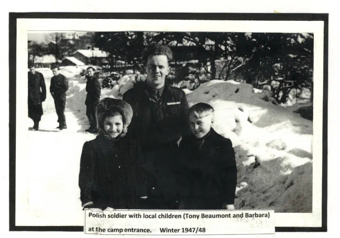 Black and white photo of a Polish soldier standing with two children, identified as Tony Beaumont and Barbara, at the entrance of a snow-covered camp. The children are smiling, and other people are visible in the background. The photo is from winter 1947/48.