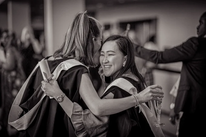 A black and white picture of two female graduates hugging in the graduation gown.