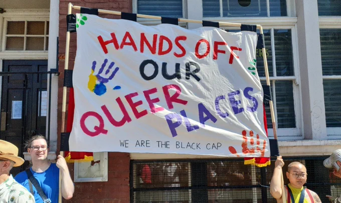 Two people holding up a large trade-union style marching banner with the slogan 'hands off our queer places' and small text saying 'we are the Black Cap'.