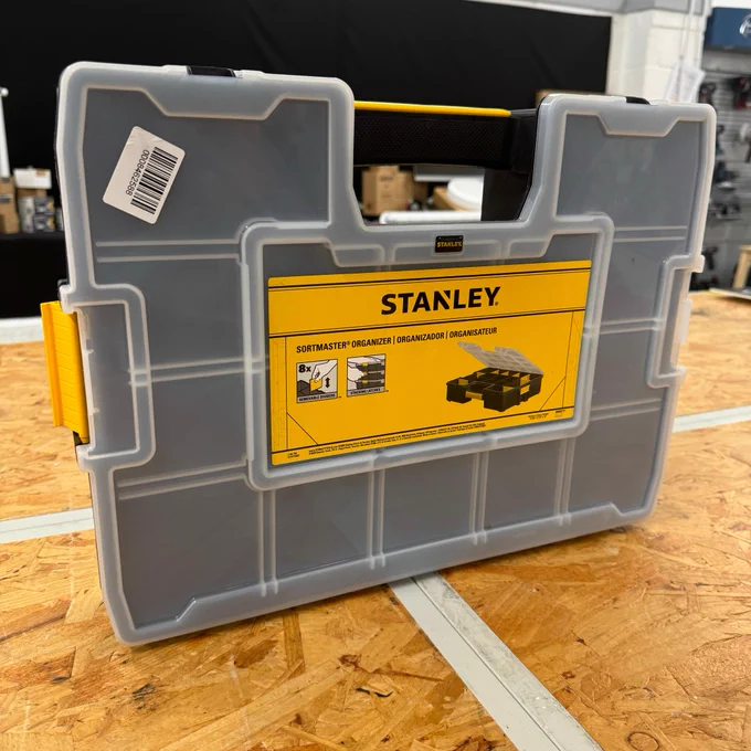 A gray Stanley brand organizer toolbox with a yellow and black label featuring an image of the product and text. The tool case is positioned on a workshop table.
