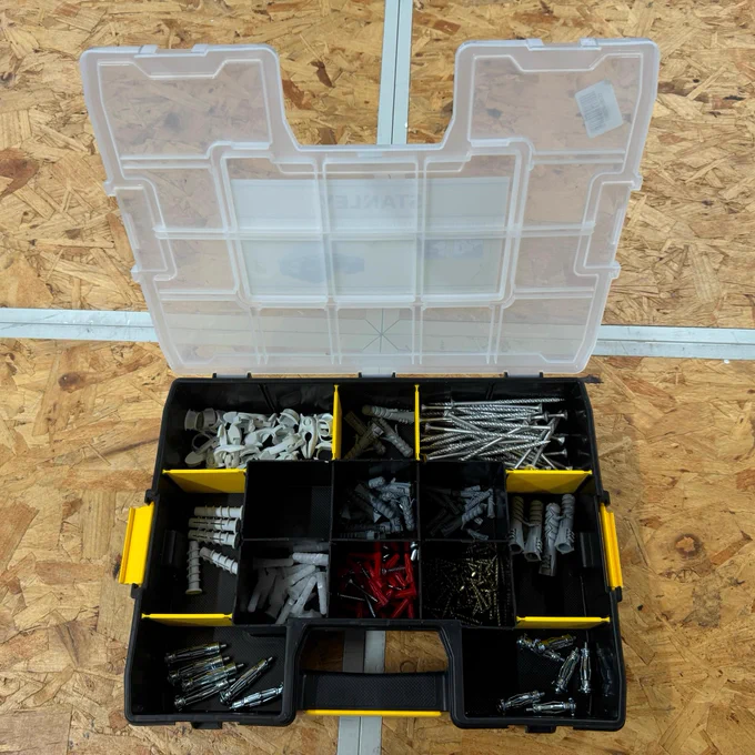An open toolbox displaying various compartments filled with nails, screws, and wall anchors, organized by type and size. The lid of the toolbox is transparent and partially raised.