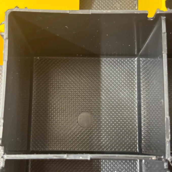 This is an image of an empty, black plastic tray with a textured interior and raised edges. The tray seems to be placed on a yellow-toned background with a patterned surface. Light reflects off some parts of the tray, creating highlights and shadows which emphasize its texture and structure.