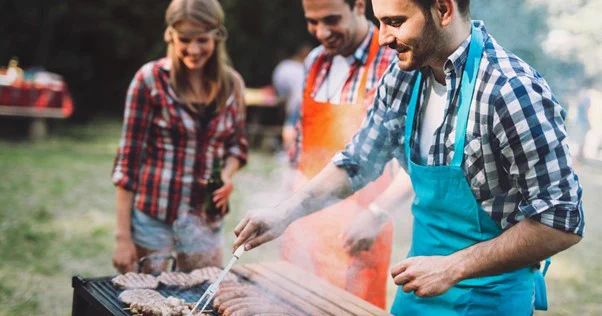 Image of man in checked shirt and blue apron is putting meat onto a barbecue, with tongs. There is meat on the barbecue and smoke coming off of it. There is another man in the background wearing a checked shirt and an orange apron. A woman is wearing a blue and black checked shirt, and everyone is smiling.