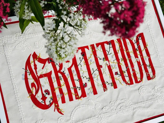 Embroidered text on cloth reads ‘Gratitudo’ with flowers woven inbetween the letters. Colourful flowers hang above