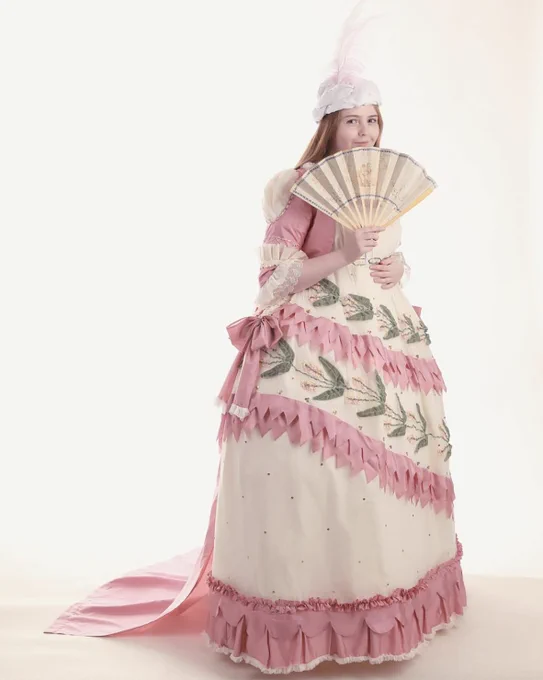A woman smiles at the camera behind her handheld fan, wearing a flowery pink 18th century dress