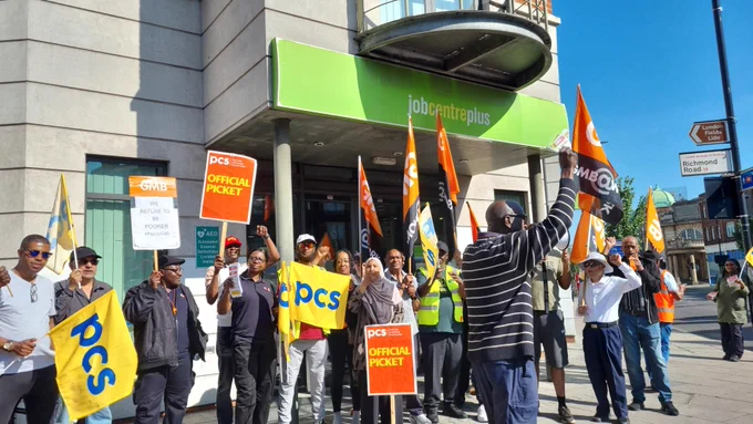 Vibrant picket line with people holding PCS flags and placards outside a "jobcentreplus" entrance.