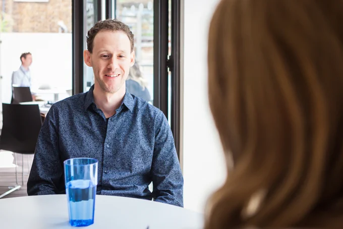 A man in blue shirt sat across a woman at a table and smiling.