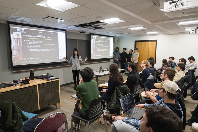 One student stands in front of a classroom full of students, a presentation up on a screen in the front of the room, which a small group of fellow students stands off to the side. The student gestures while speaking to the class. 