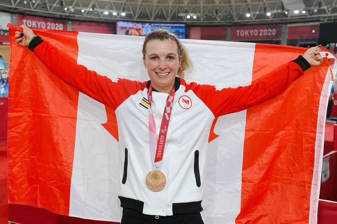 An athlete in a Team Canada uniform proudly displaying a large Canadian flag and wearing a bronze medal at the Tokyo 2020 Olympics in an indoor arena setting.
