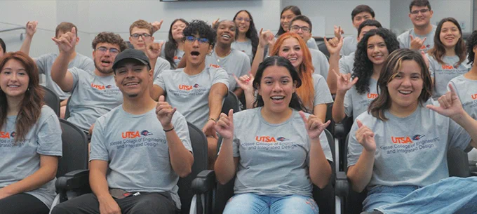 A group of enthusiastic students sitting in a classroom, all wearing gray t-shirts with "UTSA College of Engineering" printed on them. They are smiling and raising their hands in a gesture of excitement or voting.