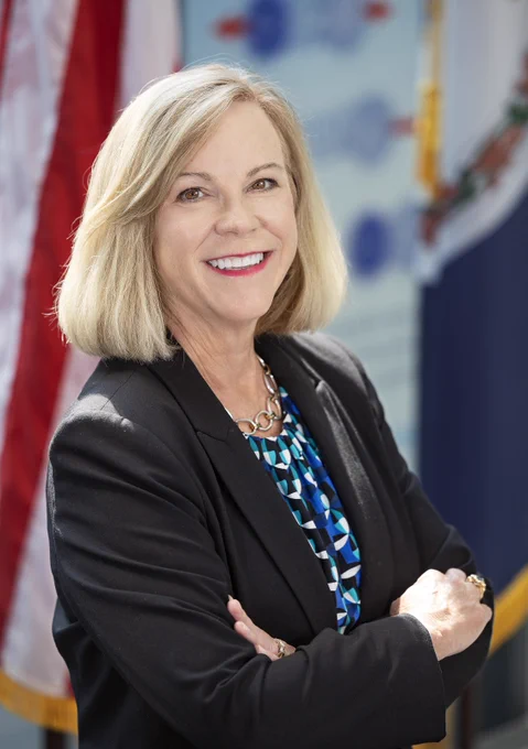 Portrait of a smiling woman with shoulder-length blonde hair, wearing a black blazer and a patterned blouse, standing in front of the American flag and another flag with seals, possibly indicating a governmental setting.