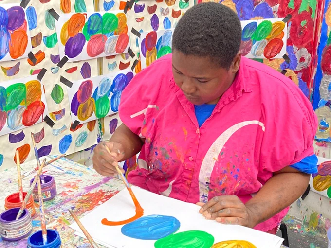 Artist Kwaga Sillingi sat at a table painting. She is surrounded by paintings of circular forms on the walls. 