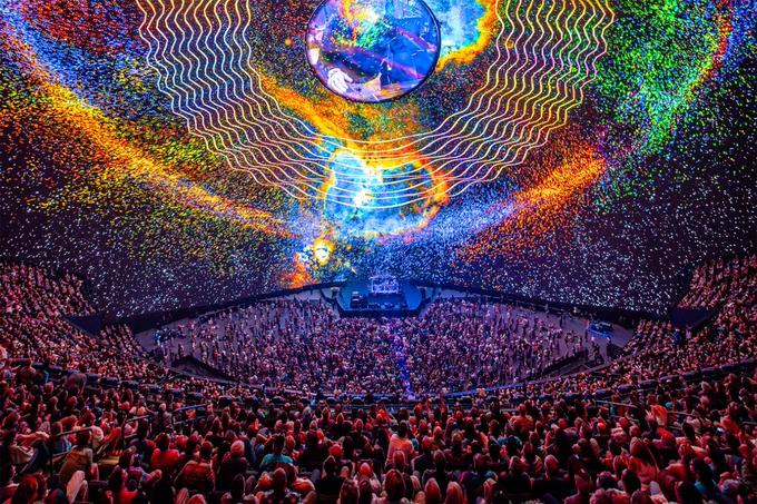 A vibrant concert scene inside an arena with a large crowd watching a spectacular light show consisting of colorful swirling patterns and a giant illuminated globe hanging above the stage.