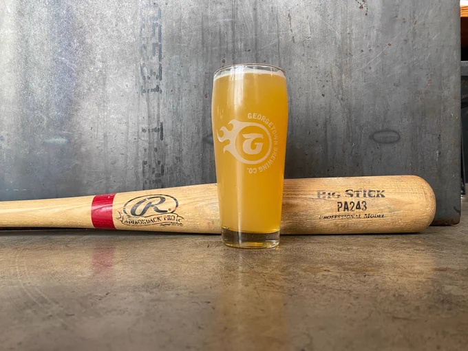 A pint of beer is positioned in front of a baseball bat that has been laid on the concrete floor. 
