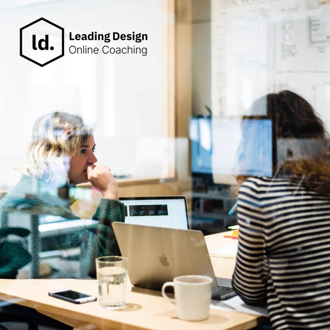 Two women engaged in a design discussion at a table with laptops and notes. The scene is set in an office environment with a whiteboard in the background. A logo in the upper left corner reads 'Leading Design Online Coaching.