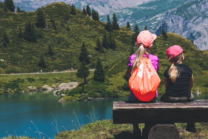 Two children wearing bright pink caps sit on a wooden bench, overlooking a serene mountain lake surrounded by lush green slopes under a clear blue sky.