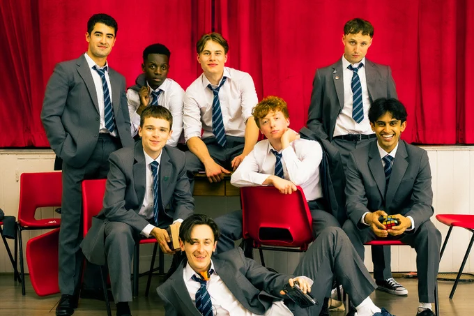A photo showing eight performers in school outfits sat in red chairs 