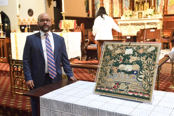 A man stands next to the sample, a woven tapestry, in front of a church altar. 