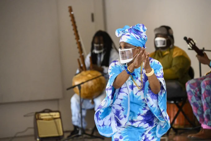 A woman in a blue boubou claps and dances while wearing a see through mask.  A band of three people with traditional west african instruments play behind her. 